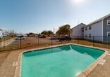 This outdoor pool area offers a welcoming recreational retreat designed for comfortable leisure and relaxation. At the heart of the space is a generously sized rectangular swimming pool with clear, turquoise water, framed by a wide concrete pool surround that provides ample room for movement and access. Adjacent to the pool, a warm-toned wooden deck serves as the primary lounging area, furnished with multiple white reclining lounge chairs positioned to take full advantage of the abundant natural sunlight. The combination of the wood deck and aggregate concrete surfaces creates a practical yet inviting poolside environment. The entire area is enclosed by a sturdy wrought-iron style perimeter fence, providing both security and a defined boundary for the space. Surrounding the pool area are multi-story residential buildings with blue and gray siding, giving the space a community-oriented feel. The open sky above allows for unobstructed sunlight throughout the day, making this an ideal setting for outdoor recreation. Mature trees are visible beyond the fence line, adding a touch of natural scenery to the surroundings. Overall, this pool area is well-suited as a shared outdoor amenity, balancing functionality with a relaxed, open-air atmosphere.