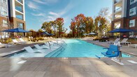 Located at 1835 Morehead Ridge Dr in Charlotte, NC, this outdoor amenity space offers a well-appointed resort-style experience within a modern residential community. The centerpiece is a generously sized swimming pool with crystal-clear blue water, framed by large-format stone pavers that extend across a spacious sun deck. The deck is furnished with contemporary white lounge chairs and chaise lounges, many of which are positioned directly within a shallow wading shelf along the pool's edge — ideal for relaxing in the water. Umbrellas in blue and red are arranged throughout the deck to provide shade, complemented by wooden pergola structures that add architectural warmth. An ADA-accessible pool lift ensures the space is welcoming to all. Surrounding the pool area, mature trees — many displaying vibrant autumn foliage — create a natural backdrop that softens the urban setting. The perimeter of the amenity area features thoughtfully designed landscaping with low shrubs, ornamental plantings, and brick retaining walls bordered by sleek metal fencing. Gated entry points with decorative ironwork provide secure, stylish access to the courtyard. The multi-story residential building rises prominently nearby, with balconies overlooking the amenity level, reinforcing the community-oriented character of this outdoor space.