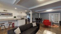 Located at 884 Thurber Dr W in Columbus, Ohio, this single-floor gathering space offers a well-designed, open-concept layout ideal for meetings, social events, or community use. The interior is defined by polished concrete floors, exposed ceiling beams, and a neutral gray and white color palette that lends a modern, inviting atmosphere throughout.

The main lounge area is generously sized, furnished with a dark sectional sofa, bold red accent chairs, and a central media console housing a wall-mounted flat-screen TV with a built-in electric fireplace beneath. A world map wall mural and decorative indoor plants add character and warmth to the space.

A secondary seating area features blue and gray upholstered armchairs arranged around a chevron-patterned area rug, with French doors opening directly to an outdoor patio — allowing natural light to flood the room and creating a seamless indoor-outdoor connection.

The kitchenette is equipped with white shaker cabinetry, a countertop microwave, a compact refrigerator, and a sink, providing practical refreshment amenities. Adjacent dining zones include both a bar-height table with stools and a standard dining table with dark chairs, offering flexible seating arrangements.

Together, these distinct areas create a cohesive, functional space well-suited for a variety of gatherings and programming needs.