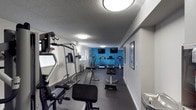 This single-floor fitness center is designed to support a comprehensive workout experience. The space is laid out efficiently, with durable dark rubberized flooring throughout that provides both safety and comfort during exercise. A bold blue accent wall at the far end of the room creates a visually energizing focal point, complemented by two wall-mounted flat-screen televisions positioned for easy viewing during cardio sessions.

The cardio section features a row of equipment including treadmills and an elliptical trainer, allowing multiple users to exercise simultaneously. On the strength training side, a multi-function cable machine and a weight bench are prominently positioned, offering a variety of resistance exercises. A utility trash receptacle is conveniently placed within the space for cleanliness and ease of use.

Overhead, flush-mount circular light fixtures provide bright, even illumination throughout the room, enhancing visibility and the overall sense of openness. A textured ceiling adds a subtle architectural detail. Natural light filters in through a window on the side wall, contributing to a pleasant and welcoming atmosphere. Framed informational or motivational prints are displayed along the walls, adding a polished, purposeful finish to this practical and functional fitness facility.