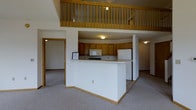 This two-story space is designed with an open-concept layout that seamlessly connects the kitchen and main living area. The kitchen is centrally positioned and features warm honey-toned wood cabinetry, a built-in microwave, electric range, and a full-size refrigerator. A white peninsula counter extends into the living space, providing a natural divide between cooking and lounging areas while maintaining an open, connected feel.

The main level is finished with neutral carpet throughout the living areas and transitions to tile flooring in the kitchen zone, offering both comfort and practicality. An additional room accessible via a wood-trimmed doorway on the main level provides flexible space for various uses.

A striking architectural highlight is the upper-level loft, which overlooks the main living area through a classic wood-railed balcony. This elevated vantage point contributes to the sense of vertical space and openness, with vaulted ceilings amplifying the airy atmosphere. The warm wood tones of the balcony railing complement the kitchen cabinetry below, tying the two levels together cohesively. Overall, the space offers a practical yet open feel, well-suited for comfortable everyday living.
