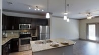 Located at 3101 Rise Dr in Morrisville, NC, this single-floor residence offers a well-appointed open-concept layout that seamlessly connects the kitchen and living areas. The kitchen is a standout feature, showcasing dark espresso cabinetry, stainless steel appliances including a refrigerator, range, and built-in microwave, complemented by granite countertops and a spacious island with a stainless steel sink — ideal for both meal preparation and casual dining. Stylish pendant lighting hangs above the island, while a track lighting system illuminates the kitchen workspace. Rich, dark hardwood-style flooring runs throughout, grounding the space with warmth and sophistication. The open living area benefits from a ceiling fan and is bathed in natural light through glass double doors that lead to an outdoor area, creating an inviting and airy atmosphere. Crown molding along the ceilings adds a refined architectural touch throughout. The overall design balances modern aesthetics with functional living, making this a comfortable and visually cohesive space.
