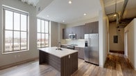 This single-floor residence showcases a cohesive modern design throughout its well-appointed spaces. The open-concept kitchen serves as a central highlight, featuring dark wood-grain cabinetry extending floor to ceiling, a white quartz countertop island with an integrated sink, and a full suite of stainless steel appliances including a French door refrigerator with ice dispenser, electric range, and built-in microwave. A white subway tile backsplash adds a clean, contemporary contrast, while wood-look plank flooring grounds the space with warmth. A large multi-pane window floods the kitchen and adjacent living area with natural light and frames urban skyline views.

The bedroom areas are finished with neutral carpet and feature tall windows that continue the unit's connection to the outdoors. One bedroom includes a ceiling fan and a spacious closet, while an additional room with wood-look flooring and an architectural cylindrical column offers versatile use. Storage is further supported by open closet systems.

The bathroom is finished with large-format light tile flooring, a dark wood-grain vanity with an integrated sink, a framed mirror, and a full soaking tub with shower. Throughout the unit, recessed lighting and a neutral color palette contribute to a clean, contemporary ambiance.