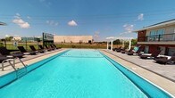 This outdoor amenity space is centered around a generously sized rectangular swimming pool with crystal-clear turquoise water, designed to serve as a focal gathering point for residents. The pool is equipped with underwater jets and is surrounded by a spacious paved deck that comfortably accommodates a row of dark-framed lounge chairs on either side, ideal for sunbathing and relaxation. A classic white pergola anchors one end of the deck, adding an architectural accent and providing a shaded retreat. The area is enclosed by a sleek black iron fence, ensuring both security and an open, airy feel. The property is identified by the Pinecrest Apartments signage visible near the entrance to the pool area. The surrounding landscape opens to broad, flat terrain under an expansive sky, allowing for abundant natural light throughout the day. The overall ambiance is clean, modern, and inviting, offering a resort-style outdoor experience. The single-level layout of the facility ensures easy, accessible flow between the pool deck and adjacent areas, making this outdoor space both functional and aesthetically appealing for everyday enjoyment.