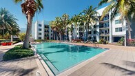 Located at 250 Congress Park Dr in Delray Beach, Florida, this outdoor amenity space offers a resort-style pool experience under the sunny South Florida sky. The centerpiece of the area is a generously sized, crystal-clear swimming pool with calm turquoise waters, framed by elegant tiling and a wide surrounding deck ideal for lounging. Rows of poolside chairs and umbrellas provide ample opportunity to relax in the Florida sunshine or find shade throughout the day. Towering palm trees are thoughtfully positioned throughout the space, adding a lush, tropical ambiance that is characteristic of the region. The pool area is nestled within a modern, multi-story building complex featuring a clean, contemporary architectural design with a white and gray exterior, large windows, and private balconies. The lush landscaping, including vibrant tropical plantings and manicured greenery, complements the modern structure and creates a welcoming, resort-like atmosphere. Under clear blue skies, this outdoor space delivers an inviting environment that blends relaxed leisure with refined design, making it an ideal gathering spot for residents and visitors seeking a true Florida outdoor lifestyle experience.