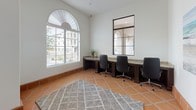 This single-floor space combines Mediterranean-inspired architecture with a practical, professional layout. Warm terracotta tiles laid in a diagonal pattern run throughout, complemented by clean white walls and rich dark wood trim that frames the doorways and windows. A striking arched fanlight window floods the workspace with natural light, offering views of the surrounding neighborhood streetscape, while horizontal blinds provide adjustable light control.

The main workspace is anchored by built-in desks with granite countertops and dark wood cabinetry, accommodating multiple workstations outfitted with ergonomic chairs — ideal for collaborative or focused work. A geometric area rug grounds the central floor space, adding texture and warmth to the room. A framed interior window with steel grid detailing connects to an adjacent area, enhancing the sense of openness.

Coastal-themed artwork, including a serene ocean scene and a bold blue-and-white striped umbrella print, adds personality and a relaxed atmosphere to the interior. A solid dark wood entry door with a glass panel leads to a connecting hallway, creating a seamless flow between spaces. The overall ambiance is calm and inviting, blending a Mediterranean aesthetic with a well-appointed, functional design suited for professional use.