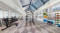 Located at 1009 Reagans Rd in Frederick, MD, this single-floor fitness center is designed to deliver a premium workout environment with thoughtful architectural detail and a well-rounded equipment offering. The space is immediately distinguished by its dramatic vaulted ceiling finished in deep navy blue with exposed beams, creating a striking visual contrast against the bright white walls. Pendant lighting hangs throughout, complementing the abundance of natural light that pours in through large floor-to-ceiling windows lining the perimeter walls, offering open views of the surrounding community.

The entrance features a bold black-framed glass door framed by a natural stone accent surround, alongside warm wood-paneled columns that add a rustic, grounded touch to the otherwise modern interior. Underfoot, a classic checkerboard-patterned carpet tile covers the entire floor, lending texture and visual interest throughout.

The fitness floor is well-equipped with a dedicated free weights area featuring a full dumbbell rack and flat benches, a multi-station cable machine, cardio equipment including treadmills and stationary bikes, a captain's chair, and a collection of stability and medicine balls. Full-length mirrors along one wall enhance the sense of space and support proper form during training. The layout is open and spacious, allowing for comfortable movement between equipment zones.