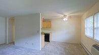 This single-floor space features an open-concept layout that connects the living area to the kitchen, creating a functional and cohesive flow. The living area is finished with neutral-toned carpeting and light gray walls, contributing to a clean and understated aesthetic. A ceiling fan with an integrated light fixture is centrally mounted, providing both air circulation and ambient lighting to the space.

The kitchen is partially separated by a half-wall partition, offering definition between the two areas while maintaining an open feel. It is equipped with warm-toned wood cabinetry, a countertop with a built-in sink, and a black dishwasher. The kitchen flooring transitions to a smooth surface, distinguishing it from the carpeted living area. The light-colored countertops complement the cabinetry, giving the kitchen a straightforward, practical design.

Throughout the space, the walls are painted in a soft, neutral tone that reflects light evenly, adding to the overall sense of openness. The layout is efficient and well-organized, making it a practical living environment. The consistent use of neutral tones across all surfaces creates a cohesive and move-in-ready atmosphere.