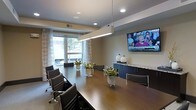 This single-floor conference room is designed to support productive professional gatherings in a polished, contemporary setting. The centerpiece is a long, dark wood conference table flanked by sleek, high-back executive chairs upholstered in black, providing ample seating for a sizable group. A linear pendant light fixture runs above the table, complementing the recessed ceiling lighting to create a well-lit, focused atmosphere.

A large wall-mounted flat-screen television serves as a prominent feature on one wall, making the space well-suited for presentations and collaborative discussions. Adjacent to the screen, dark cabinetry with a granite-style countertop provides functional storage and display space, accented by decorative ceramic vessels.

Natural light filters into the room through a window dressed with sheer curtains, offering a soft connection to the outdoors while maintaining privacy. Potted greenery placed along the table adds a refreshing touch, softening the room's professional aesthetic. Neutral wall tones and carpeted flooring contribute to a clean, understated ambiance that keeps the focus on functionality and comfort.