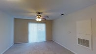 This single-floor space offers a straightforward and functional layout designed for comfortable everyday living. The main living area is finished with wall-to-wall carpet in a neutral tone, providing a warm and inviting foundation throughout. A ceiling fan with an integrated light fixture is centrally mounted, ensuring consistent air circulation and ambient lighting. A large sliding glass door, dressed with vertical blinds, serves as the room's primary source of natural light, brightening the interior and offering potential access to an outdoor area. The walls are painted in a soft, neutral hue that complements the light carpeting, creating a cohesive and understated aesthetic. Recessed HVAC vents are visible in the ceiling, indicating a built-in climate control system. Clean white baseboards frame the perimeter of the room, adding a crisp finishing detail. The textured ceiling adds subtle visual interest overhead. Overall, the space presents a clean, move-in ready environment with practical features and a calm, neutral palette that provides a versatile backdrop for a variety of interior styles and furnishing preferences.