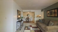 This single-floor residence offers a thoughtfully designed open-concept layout that seamlessly connects the living, dining, and kitchen areas. The living room is a welcoming space, featuring plush carpeting, a geometric area rug in warm earth tones, and ample room for comfortable seating. A decorative console table with ornate metalwork and an accent lamp adds character to the space. The living area flows naturally into the dining room, which is distinguished by a tray ceiling detail and hardwood-style flooring, lending an elegant sense of definition to the space. A round dining table with classic chairs sits beneath a circular wall mirror, creating an intimate and stylish setting. Just beyond, the kitchen is visible through a pass-through window, featuring light wood cabinetry and a clean, functional design. A textured popcorn ceiling adds a classic touch throughout the shared living spaces. Neutral wall tones, including a tasteful accent wall in deep gray in the living area, provide a versatile backdrop that complements the warm, earthy furnishings. The layout is efficient and cohesive, making excellent use of the available space while maintaining a comfortable and inviting ambiance throughout.