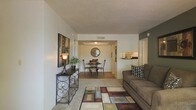 This single-floor residence offers a thoughtfully designed open-concept layout that seamlessly connects the living, dining, and kitchen areas. The living room is a welcoming space, featuring plush carpeting, a geometric area rug in warm earth tones, and ample room for comfortable seating. A decorative console table with ornate metalwork and an accent lamp adds character to the space. The living area flows naturally into the dining room, which is distinguished by a tray ceiling detail and hardwood-style flooring, lending an elegant sense of definition to the space. A round dining table with classic chairs sits beneath a circular wall mirror, creating an intimate and stylish setting. Just beyond, the kitchen is visible through a pass-through window, featuring light wood cabinetry and a clean, functional design. A textured popcorn ceiling adds a classic touch throughout the shared living spaces. Neutral wall tones, including a tasteful accent wall in deep gray in the living area, provide a versatile backdrop that complements the warm, earthy furnishings. The layout is efficient and cohesive, making excellent use of the available space while maintaining a comfortable and inviting ambiance throughout.