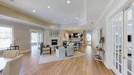 Located at 14411 Calloway Gap Rd in Raleigh, NC, this single-floor space offers a thoughtfully designed interior that balances comfort and sophistication. The main lounge area is anchored by a classic white fireplace, serving as a focal point beneath a gallery of framed botanical prints. A tray ceiling adds architectural dimension to the room, while recessed lighting and elegant pendant fixtures ensure the space is well-lit throughout the day and evening. Smooth hardwood flooring extends across the entire area, lending warmth and cohesion to the open layout. Generous seating arrangements — including upholstered sofas, accent chairs, and a coffee table — invite relaxed gathering and conversation. Toward the rear of the space, a counter-height bar area with pendant lighting provides an additional social setting, ideal for casual interaction. Large windows and glass-paneled doors on multiple sides flood the interior with natural light and offer views of the surrounding landscape. The neutral color palette of soft grays, warm whites, and natural wood tones creates a refined yet welcoming ambiance. This well-appointed space is designed to accommodate a variety of uses, from quiet relaxation to community gatherings.