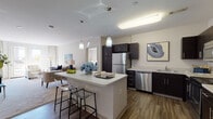 Located at 100 Exchange St in Malden, Massachusetts, this well-appointed single-floor residence offers a thoughtfully designed open-concept layout with modern finishes throughout. The kitchen serves as a central hub, featuring dark espresso cabinetry, light quartz countertops, stainless steel appliances including a refrigerator, dishwasher, microwave, and range, along with a center island with bar seating and pendant lighting. Rich hardwood-style flooring grounds the kitchen and flows into the adjacent dining area. The living room is bright and welcoming, with plush carpeting, a wall-mounted TV, and French doors that open to a private outdoor balcony, allowing abundant natural light and urban views. The primary bedroom is generously sized with multiple windows, soft carpeting, and direct access to a full bathroom featuring a dark-cabinet vanity with a white countertop and a shower-tub combination. A second bedroom offers flexible use as a guest room or home office, complete with a daybed, workspace, and a large window with street views. The second bathroom mirrors the same clean, contemporary aesthetic with a single-sink vanity and walk-in shower. Throughout the unit, neutral tones and consistent dark wood accents create a cohesive, modern ambiance.