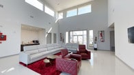 This single-floor space makes an immediate impression with its dramatic double-height ceilings and expansive wall of windows that flood the interior with natural light. The main living area is defined by clean architectural lines, with crisp white walls and high clerestory windows that draw the eye upward, enhancing the sense of openness and volume.

The lounge area is furnished with a large, contemporary white sectional sofa anchored by a rich red area rug, which introduces a striking contrast against the neutral palette. Complementing the sectional are deep burgundy leather armchairs and an ottoman, adding warmth and a bold design element to the space. A set of round wooden coffee tables grounds the seating arrangement.

Abstract artwork displayed on the walls adds visual interest and reinforces the modern aesthetic. Near the back wall, a small beverage station provides functional convenience. The light-toned flooring reflects natural light throughout, further amplifying the airy atmosphere. Large exterior-facing windows offer views of the surrounding environment, seamlessly connecting the interior to the outdoors.