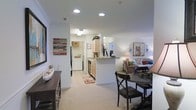 This single-floor residence is designed with an open-concept layout that connects the kitchen, dining, and living areas into one cohesive and functional space. The kitchen features light wood cabinetry, a peninsula with breakfast bar seating, and stainless steel appliances. Recessed lighting illuminates the space throughout, complementing the neutral color palette of white walls and light-toned carpeting that extends through the living and dining areas. The dining space accommodates a round table with cross-back chairs, offering an intimate setting adjacent to the kitchen. The living area, visible from the kitchen, is warmly furnished and benefits from the open sightlines created by the flowing layout. A dedicated in-unit laundry area with a stacked or side-by-side washer and dryer is conveniently tucked near the kitchen, adding practical functionality. Decorative wall art and thoughtful furnishings contribute to the welcoming ambiance. Crown molding and recessed lighting add subtle architectural detail, while the overall design emphasizes ease of movement and a comfortable, livable atmosphere throughout the home.