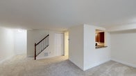 This two-floor space is designed with a clean, functional layout that balances openness with defined areas. The main level features a spacious living area finished with plush light-colored carpeting, creating a comfortable and neutral foundation. Crisp white walls and a smooth white ceiling contribute to a bright, airy atmosphere throughout.

A staircase with warm wood handrails and white balusters leads to the upper floor, serving as a subtle architectural focal point within the living space. Nearby, a closeted storage area and an additional doorway provide convenient access to adjacent spaces.

A pass-through opening in the partial wall connects the living area to the kitchen, offering a glimpse of rich, dark wood cabinetry and stone countertops. This design element promotes an open feel between the two spaces while maintaining visual separation. The kitchen appears well-appointed with ample cabinetry for storage.

A floor vent is integrated into the baseboard area, indicating a forced-air climate system. Overall, the space presents a neutral, move-in-ready aesthetic with practical layout choices that support comfortable everyday living across both floors.