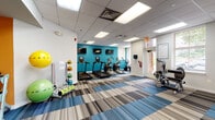 This fitness center is designed to accommodate a comprehensive range of workout routines within a single, open-plan space. The interior is visually energized by bold accent walls in teal and orange, contrasting against neutral white walls and a drop-panel ceiling fitted with recessed fluorescent lighting. The striped carpet tiles in blue, gray, and beige tones add a modern, dynamic quality to the floor throughout the facility.

The cardio zone features a row of treadmills and elliptical machines, each equipped with individual display screens. Multiple wall-mounted televisions are positioned along the teal accent wall, providing entertainment during workouts. A cable rowing machine and additional cardio equipment round out this section.

The strength-training area houses a multi-station cable machine by Hoist, a seated weight machine, and an adjustable workout bench paired with a tiered dumbbell rack. A dedicated functional training nook, highlighted by the orange accent wall, offers stability balls, medicine balls, resistance bands, and foam rollers for flexibility and cross-training exercises.

Natural light enters through multiple windows fitted with blinds, and direct exterior access is provided via a ground-level door, enhancing ventilation and convenience throughout the space.