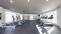 This fitness center is designed to provide a comfortable and functional workout environment. The space is generously sized, with a clean, open layout that allows for easy movement between equipment. Along both sides of the room, a lineup of cardio machines—including treadmills, ellipticals, and stair climbers—are neatly arranged to maximize the available floor space. The flooring consists of durable rubber gym matting, providing both safety and sound absorption during workouts.

The ceiling is finished in a neutral gray tone and outfitted with multiple ceiling fans, ensuring consistent air circulation throughout the room. Recessed lighting supplements the abundant natural light that streams in through large windows positioned along the perimeter walls, creating a bright and energizing atmosphere. Mirrors along the interior walls enhance the sense of space while allowing users to monitor their form during exercise. A wall-mounted television is also visible, offering entertainment during cardio sessions.

The overall ambiance is clean, modern, and functional, with a neutral white and gray color palette that contributes to a focused and motivating workout environment.