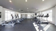 This fitness center is designed to provide a comfortable and functional workout environment. The space is generously sized, with a clean, open layout that allows for easy movement between equipment. Along both sides of the room, a lineup of cardio machines—including treadmills, ellipticals, and stair climbers—are neatly arranged to maximize the available floor space. The flooring consists of durable rubber gym matting, providing both safety and sound absorption during workouts.

The ceiling is finished in a neutral gray tone and outfitted with multiple ceiling fans, ensuring consistent air circulation throughout the room. Recessed lighting supplements the abundant natural light that streams in through large windows positioned along the perimeter walls, creating a bright and energizing atmosphere. Mirrors along the interior walls enhance the sense of space while allowing users to monitor their form during exercise. A wall-mounted television is also visible, offering entertainment during cardio sessions.

The overall ambiance is clean, modern, and functional, with a neutral white and gray color palette that contributes to a focused and motivating workout environment.
