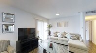 This single-floor residence at Deer Creek offers a contemporary living environment defined by clean lines and a light, neutral palette. The living area serves as the focal point of the space, featuring wood-style flooring that adds warmth underfoot while complementing the crisp white walls and ceiling. Recessed lighting provides consistent illumination throughout, supplemented by generous natural light streaming in through a large sliding glass door fitted with sheer white curtains. This glazed access point draws the eye and connects the interior to the outdoors, creating an airy, open atmosphere. The room comfortably accommodates a sectional sofa with coordinating accent pillows, a glass-top coffee table, and a media console, demonstrating a practical and well-proportioned layout. Decorative touches, including potted tropical plants and framed wall art, add character without overwhelming the space. The overall ambiance is calm and contemporary, with a neutral color scheme that provides a versatile backdrop suitable for a range of furnishing styles. The cohesive design and thoughtful use of natural light make this a comfortable and inviting single-level living space.