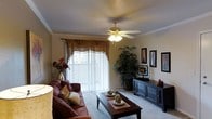 This single-floor residence centers around a warmly appointed living room that combines classic design elements with comfortable functionality. Crown molding frames the ceiling, adding an elegant architectural detail, while a ceiling fan with an integrated light fixture provides both airflow and ambient illumination. A large sliding glass door, dressed with decorative valance draping and vertical blinds, allows natural light to filter into the space and serves as a focal point along one wall.

The living room is furnished with a rich, leather-toned sofa accented with decorative throw pillows, anchored by a dark wood coffee table that features a lower display shelf. A matching dark wood media console along the opposite wall offers ample storage through its cabinet and shelf configuration, suitable for housing entertainment components. Framed wall art is arranged thoughtfully above the console, contributing to the room's curated aesthetic.

Lush indoor greenery, including a tall potted plant in the corner, introduces a natural, organic element that enhances the overall warmth of the space. Neutral carpeting throughout ties the room together, creating a cohesive and inviting atmosphere well-suited to everyday living.