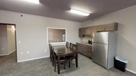 This single-floor unit is designed with a practical, straightforward layout that combines the kitchen and dining areas into one cohesive space. The kitchen is equipped with medium-toned wood cabinetry, a stainless steel refrigerator, a countertop microwave, and a sink, providing essential functionality for everyday use. A pass-through window connects the kitchen area to an adjacent space, adding an open, communicative feel between rooms. The dining area accommodates a dark wood rectangular table with seating for six, positioned beneath flush-mounted fluorescent lighting that illuminates the space efficiently. Textured ceilings and neutral-toned walls contribute to the understated aesthetic throughout. Patterned carpet flooring extends across the main living and dining areas, adding a layer of texture underfoot. A wall-mounted flat-screen television is positioned opposite the kitchen, offering a clear sightline from the dining and living zones. Closet spaces feature white wire shelving systems, maximizing vertical storage capacity. Horizontal window blinds provide light control and privacy. Overall, the unit presents a clean, functional environment suited for comfortable day-to-day living.