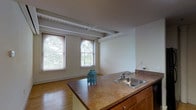 This two-story space showcases a thoughtful layout that combines functional design with character-rich architectural details. The kitchen serves as a central gathering point, featuring a wraparound island with a built-in stainless steel sink and warm wood cabinetry beneath a countertop surface. The open layout connects the kitchen seamlessly to the adjoining living area, creating a cohesive, flowing space ideal for everyday living.

Overhead, exposed beams and ductwork add an industrial character to the otherwise neutral interior, lending the space a loft-like quality. The ceilings are generously proportioned, enhancing the sense of openness. Hardwood flooring runs throughout, adding warmth and continuity between areas.

Natural light is a defining feature of this space, with multiple windows — including an arched accent window — allowing daylight to pour in and frame views of mature trees outside. Light-toned walls throughout complement the natural brightness.

A hallway connects the main living areas, with light switches and subtle architectural details visible along the corridor. The overall ambiance is bright, open, and functional, with a mix of classic and industrial design elements that give the space a distinctive character.