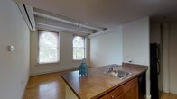 This two-story space showcases a thoughtful layout that combines functional design with character-rich architectural details. The kitchen serves as a central gathering point, featuring a wraparound island with a built-in stainless steel sink and warm wood cabinetry beneath a countertop surface. The open layout connects the kitchen seamlessly to the adjoining living area, creating a cohesive, flowing space ideal for everyday living.

Overhead, exposed beams and ductwork add an industrial character to the otherwise neutral interior, lending the space a loft-like quality. The ceilings are generously proportioned, enhancing the sense of openness. Hardwood flooring runs throughout, adding warmth and continuity between areas.

Natural light is a defining feature of this space, with multiple windows — including an arched accent window — allowing daylight to pour in and frame views of mature trees outside. Light-toned walls throughout complement the natural brightness.

A hallway connects the main living areas, with light switches and subtle architectural details visible along the corridor. The overall ambiance is bright, open, and functional, with a mix of classic and industrial design elements that give the space a distinctive character.