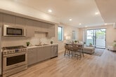 Located at 11100 Hartsook St in Los Angeles, CA, this single-floor unit showcases a contemporary design with a seamless open-concept layout connecting the kitchen, dining, and living areas. Light wood-style flooring runs throughout, contributing to a bright and cohesive aesthetic.

The kitchen is well-appointed with gray shaker-style cabinetry extending to the ceiling, white quartz countertops, and a full suite of stainless steel appliances including a gas range, over-the-range microwave, refrigerator, and dishwasher. A stainless steel sink with a modern gooseneck faucet completes the space.

The dining area accommodates a bar-height table with seating and sits adjacent to both the kitchen and the living room, facilitating easy flow between spaces. The living room is generously sized, featuring recessed lighting and a large sliding glass door that opens to a private balcony with views of surrounding greenery, bringing in ample natural light.

The bathroom features a wood-toned vanity with a quartz countertop, round mirror, white penny-tile flooring, and a tiled tub-shower combination with sliding glass doors. Neutral tones and recessed lighting give the bathroom a clean, modern finish throughout.