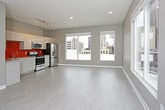 This contemporary single-floor unit showcases a clean, modern design with a neutral gray-toned palette, light wood-look flooring, and crisp white baseboards that create a cohesive and polished aesthetic throughout. The open-concept living and kitchen area serves as the heart of the space, with an abundance of natural light streaming in through large sliding windows that frame urban cityscape views. The kitchen is a standout feature, boasting sleek white flat-front cabinetry extending to the ceiling, granite-look countertops, a striking orange glass tile backsplash, a stainless steel sink, and a full suite of stainless steel appliances including a range, over-the-range microwave, and French door refrigerator. Track lighting overhead provides focused illumination across the kitchen. The living area is generously proportioned and open, with recessed lighting and additional windows offering further city views and ample natural light. A dedicated room, suitable as a bedroom or flexible space, features recessed lighting and its own HVAC vent. A spacious walk-in closet is outfitted with multi-tiered wire shelving and features dark carpeting for a practical touch. The full bathroom includes a combination soaking tub and shower with a curved rod, a granite-top vanity with dark wood cabinetry, and light tile flooring, completing this well-appointed urban unit.