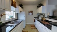 This single-floor residence offers a well-organized layout designed for practical, comfortable living. The hallway serves as the central connector of the home, finished with neutral carpet and illuminated by a flush-mount ceiling light. A standout feature is the dedicated in-unit laundry closet tucked neatly into the hallway, housing a full-sized washer and dryer behind bifold doors — a highly convenient addition to everyday living.

The living room is a welcoming space featuring plush carpet, a textured ceiling, and a large sliding glass door dressed with vertical blinds that allow generous natural light to fill the room. The area comfortably accommodates seating and provides a relaxed, cozy ambiance.

The galley-style kitchen is efficiently designed with white cabinetry extending to the ceiling, dark granite-look countertops, and a double stainless steel sink positioned beneath a window. White appliances, including a refrigerator and electric range, complement the checkered tile backsplash. Tile flooring runs throughout the kitchen, and upper shelving provides ample storage. The overall aesthetic is clean, classic, and highly functional.