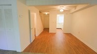This two-story home offers a straightforward, functional layout with a clean and bright interior. The main level is anchored by an open living area finished with warm hardwood-style flooring that extends throughout the space, lending a cohesive and inviting feel underfoot. A ceiling fan is centrally mounted overhead, providing both light and air circulation to the main living zone.

A structural beam subtly defines the transition between areas, adding a modest architectural detail to the otherwise smooth, white-painted walls and ceilings. The open sightline from the living area leads naturally toward a dedicated kitchen space, which is visible at the rear and features white cabinetry, a window that allows natural light to filter in, and what appears to be standard kitchen appliances.

A staircase situated toward the back of the main level provides access to the second floor, accompanied by a door nearby that likely leads to a utility or storage area. An additional door on the left side of the living space offers further room access. The overall ambiance is neutral and well-lit, presenting a practical and move-in-ready environment throughout both levels.