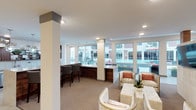 Located in Dallas, this single-floor community lounge space offers a well-appointed and inviting environment designed for both relaxation and socializing. The layout is thoughtfully divided into distinct functional zones that flow seamlessly into one another. A built-in bar area with a rich dark wood counter and high-back bar seating provides an ideal spot for casual gatherings, while pendant lighting adds a refined touch overhead. Adjacent to the bar, a lounge area features elegant cream and gold accent chairs paired with a clean-lined coffee table, creating a comfortable conversational setting. Warm neutral tones throughout the carpeted flooring and white walls lend the space a polished, contemporary aesthetic. Recessed lighting illuminates the interior evenly, complementing the abundant natural light that pours in through the floor-to-ceiling windows lining the exterior wall. These expansive windows frame picturesque views of a resort-style outdoor pool and surrounding courtyard, seamlessly connecting the interior to the outdoor amenity space. Structural columns are integrated throughout in a clean, minimal fashion, adding architectural interest without interrupting the open feel of the room. Overall, the space strikes a balance between sophistication and comfort, making it a versatile community hub.