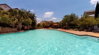 This outdoor pool area serves as a well-appointed recreational amenity within a multi-story residential community. The centerpiece is a generously sized swimming pool with crystal-clear turquoise water, complemented by a separate hot tub situated directly adjacent. The surrounding sundeck is finished in a durable concrete surface and is furnished with multiple lounge chairs and chaise lounges, providing ample space for relaxation and sunbathing under open skies.

A shaded gazebo anchors one end of the pool deck, offering a covered retreat from direct sun, while mature trees — including palms and leafy shade trees — are planted throughout the perimeter, creating a lush, resort-like atmosphere. The pool area is enclosed and accessed through a dedicated entrance featuring a covered entryway with a posted pool rules sign, dark wood-framed glass doors, and an exterior wall sconce.

Views from the pool deck look out toward the surrounding multi-story residential buildings, which feature brick and wood siding exteriors with private balconies overlooking the amenity. The entire space benefits from abundant natural sunlight throughout the day, with open sky views that enhance the outdoor experience. The thoughtful layout and landscaping combine to create a functional and inviting outdoor leisure environment.