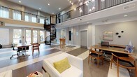 This single-floor space makes a striking first impression with its grand, double-height lobby anchored by a dramatic spiral staircase and an upper mezzanine level framed by sleek black railings. A cascading branch-style chandelier draws the eye upward, emphasizing the soaring ceiling height. Large windows allow natural light to fill the open floor plan, which features polished tile flooring in a two-tone pattern and neutral wall tones throughout.

The lounge area is furnished with crisp white sofas accented by chartreuse throw pillows, complemented by cowhide ottomans and a sleek arc floor lamp. Floor-to-ceiling mirrored panels add depth and visual interest to this seating zone, creating an upscale, modern ambiance.

A dedicated meeting or dining area features a solid wood table surrounded by cream upholstered chairs with nail-head trim, a wall-mounted flat-screen television, and soft sage-green walls decorated with curated artwork and sculptural wall accents.

The kitchenette is thoughtfully appointed with light granite countertops, white shaker-style cabinetry, a double stainless steel sink, and a full white subway tile backsplash, providing practical amenities within the space.