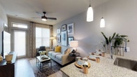 This single-floor residence showcases a thoughtfully designed open-concept layout that connects the dining and living spaces in a seamless, functional flow. The kitchen peninsula features striking granite countertops and is illuminated by elegant pendant lights, making it a natural gathering point. The dining area is set beneath the warm glow of pendant lighting, offering a polished yet comfortable setting for meals and entertaining.

The living area is anchored by a contemporary ceiling fan with a sleek dark finish, complementing the neutral grey walls and light hardwood flooring that run throughout the space. A stylishly arranged gallery wall adds a personalized, curated touch above the sofa, which is adorned with coordinating accent pillows in muted tones and a pop of mustard yellow. Natural light filters into the room through a large window dressed with layered curtains, enhancing the bright and airy ambiance.

The overall aesthetic is modern and cohesive, blending clean lines with warm textures and metallic accents. The space feels open and well-proportioned, offering a comfortable and visually appealing environment suited for both everyday living and entertaining.