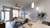 This single-floor residence showcases a thoughtfully designed open-concept layout that connects the dining and living spaces in a seamless, functional flow. The kitchen peninsula features striking granite countertops and is illuminated by elegant pendant lights, making it a natural gathering point. The dining area is set beneath the warm glow of pendant lighting, offering a polished yet comfortable setting for meals and entertaining.

The living area is anchored by a contemporary ceiling fan with a sleek dark finish, complementing the neutral grey walls and light hardwood flooring that run throughout the space. A stylishly arranged gallery wall adds a personalized, curated touch above the sofa, which is adorned with coordinating accent pillows in muted tones and a pop of mustard yellow. Natural light filters into the room through a large window dressed with layered curtains, enhancing the bright and airy ambiance.

The overall aesthetic is modern and cohesive, blending clean lines with warm textures and metallic accents. The space feels open and well-proportioned, offering a comfortable and visually appealing environment suited for both everyday living and entertaining.