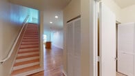 This two-story residence is thoughtfully laid out across two levels, connected by a staircase visible from the main entry corridor. The ground floor features an open-concept kitchen and living area, where warm hardwood-style flooring flows continuously throughout. The kitchen is well-appointed with honey-toned cabinetry, dark granite countertops, a curved breakfast bar island, and a full suite of stainless steel appliances including a refrigerator with ice maker, range, and built-in microwave. A pendant light fixture adds a focal point to the adjoining dining and living space, which benefits from recessed lighting and a window view toward the exterior. A hallway closet with louvered bifold doors provides convenient main-level storage. Upstairs, an open loft-style living space is anchored by a ceiling fan, natural light from a window, and a balcony-style railing overlooking the lower level. This floor also includes a spacious walk-in closet with wire shelving, a full bathroom featuring both a soaking tub and a separate glass-enclosed shower with tile surround, and an additional bathroom with a vanity and louvered storage closet. Light, neutral tones are consistent throughout.