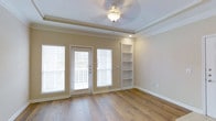 This single-floor space combines classic architectural detailing with practical design elements to create a warm and inviting environment. The main living area is anchored by rich, dark hardwood flooring that runs throughout, adding warmth and character to the space. A distinctive tray ceiling with crown molding lends an elevated sense of height and architectural refinement, while a decorative ceiling light fixture serves as a subtle focal point overhead.

Natural light flows generously into the room through a glass-panel exterior door flanked by large windows fitted with horizontal blinds, offering both brightness and privacy control. The door provides direct access to an outdoor balcony, extending the living space and offering outdoor views. Adjacent to the entry point, a set of built-in open shelving units is seamlessly integrated into the wall, providing functional display and storage space while adding a custom, finished look to the room.

The neutral, warm-toned walls complement the hardwood floors and white trim, creating a cohesive and versatile palette suitable for a variety of interior styles. Recessed lighting supplements the natural light, ensuring the space remains well-lit throughout the day. The overall ambiance is clean, airy, and move-in ready, with thoughtful details that reflect quality craftsmanship throughout.