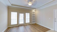This single-floor space combines classic architectural detailing with practical design elements to create a warm and inviting environment. The main living area is anchored by rich, dark hardwood flooring that runs throughout, adding warmth and character to the space. A distinctive tray ceiling with crown molding lends an elevated sense of height and architectural refinement, while a decorative ceiling light fixture serves as a subtle focal point overhead.

Natural light flows generously into the room through a glass-panel exterior door flanked by large windows fitted with horizontal blinds, offering both brightness and privacy control. The door provides direct access to an outdoor balcony, extending the living space and offering outdoor views. Adjacent to the entry point, a set of built-in open shelving units is seamlessly integrated into the wall, providing functional display and storage space while adding a custom, finished look to the room.

The neutral, warm-toned walls complement the hardwood floors and white trim, creating a cohesive and versatile palette suitable for a variety of interior styles. Recessed lighting supplements the natural light, ensuring the space remains well-lit throughout the day. The overall ambiance is clean, airy, and move-in ready, with thoughtful details that reflect quality craftsmanship throughout.