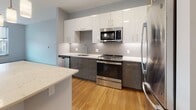 Located at 100 Fountain St in downtown Providence, Rhode Island, this single-floor unit showcases a clean, contemporary design throughout. The open-concept living and kitchen area is anchored by warm hardwood flooring and neutral gray walls, creating a bright and cohesive atmosphere. Pendant lighting hangs above a central kitchen island with a quartz-style countertop, offering both workspace and casual seating.

The kitchen is well-appointed with two-tone cabinetry—white upper cabinets and gray lower cabinets—paired with a textured tile backsplash and stainless steel appliances, including a range, microwave, dishwasher, and refrigerator. A second quartz countertop surface extends along the perimeter, maximizing prep space.

The bedroom features plush carpeting and dual windows that bring in natural light and urban views of the surrounding Providence streetscape, contributing to an airy feel.

The bathroom is finished in a modern, minimalist style with light tile flooring, subway-tiled walk-in shower with a glass door, a floating vanity with a white countertop, and a stacked washer/dryer unit conveniently integrated into the space. Overall, the unit presents a polished, functional living environment.
