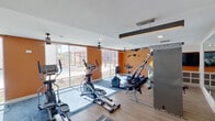 This fitness center is a comprehensively equipped workout facility designed to support a wide range of exercise routines. The space is defined by its vibrant burnt-orange accent walls, which contrast boldly against clean white ceilings and light hardwood-style flooring, creating an energetic and motivating atmosphere. Large floor-to-ceiling windows line one side of the room, flooding the interior with natural light and offering open views of the surrounding exterior, enhancing the sense of space throughout.

The cardio zone is well-stocked with multiple elliptical trainers, stationary bikes—including upright and recumbent models—and treadmills, providing ample options for cardiovascular training. A dedicated strength training area features a multi-station cable machine, a dumbbell rack with a full range of free weights, and various resistance equipment. Stability and flexibility training are also supported, with exercise balls and additional accessories neatly stored along the walls.

Ceiling fans are installed throughout to promote air circulation, complementing the recessed lighting that ensures even illumination across the entire floor. A wall-mounted television adds an entertainment option for users during workouts. The mix of rubber-bordered area rugs and smooth flooring helps define different workout zones while offering comfort and safety underfoot. Overall, this fitness center is a functional, modern, and well-organized facility built to serve diverse fitness needs.