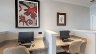 Located at 5407 Edwards Plantation Dr in Columbus, Ohio, this single-floor business center offers a focused and functional environment designed to support productivity. The space features multiple dedicated computer workstations, each equipped with a monitor, keyboard, and mouse, thoughtfully arranged to provide individual privacy through a centrally positioned white partition divider. The workstation surfaces are finished in a warm neutral tone, complemented by cushioned task chairs that ensure comfort during extended use. The walls are painted in a clean, light neutral hue that enhances the sense of openness and professionalism throughout the space. Decorative framed artwork adorns the walls, including a bold, large-scale piece featuring red leaf motifs and a smaller botanical-style print in a dark wood frame, adding warmth and visual interest to the otherwise utilitarian setting. The flooring is finished in a neutral, low-pile material that is both durable and practical for a high-use environment. Recessed and ambient lighting provides consistent, even illumination across the workspace. Overall, the space strikes a balance between professional functionality and inviting décor, making it a well-appointed setting for focused work, research, or administrative tasks.
