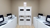 This shared laundry facility is designed to be both functional and comfortable for residents. The space is laid out across a single level and accessed through elegant French doors with frosted glass panels, lending a polished touch to an everyday utility space. The flooring throughout consists of light-toned ceramic tiles, contributing to a clean and low-maintenance environment.

The laundry area is equipped with multiple top-loading washing machines and stacked washer-dryer units, providing ample capacity for shared use. A dedicated folding station features a white countertop with cabinet storage below, offering a convenient and organized surface for laundry tasks. A freestanding garment rack is also available for hanging items.

A separate seating and waiting area allows residents to relax while their laundry cycles run. This section includes a dark wood table with seating, a wall-mounted flat-screen television, and decorative artwork depicting city street scenes, adding visual warmth to the neutral taupe walls. Overhead fluorescent lighting ensures the entire space is well-illuminated throughout. Informational notices are posted on the walls, keeping usage guidelines clearly visible to all users.