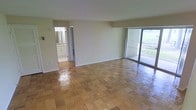 This single-floor space offers an open and airy layout defined by its distinctive parquet wood flooring, which adds warmth and visual texture throughout the main living area. The floors are finished in a classic block pattern, lending a timeless character to the space. Large sliding glass doors dominate one wall, flooding the interior with natural light and providing direct views of a green outdoor area, creating a seamless connection between indoors and out. The exterior brick wall visible through the glass adds an architectural element of interest. The neutral, cream-toned walls keep the space feeling bright and versatile. An adjacent room, visible through an open doorway, appears to be a bathroom or utility area, complete with cabinetry and its own lighting fixture, adding functional convenience to the layout. The open floor plan allows for flexible use of the main living space, accommodating a variety of furniture arrangements. Overall, the space presents a clean, well-lit environment with practical features and a straightforward design that balances functionality with comfortable, everyday living.