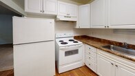 Located at 26 Yorkshire Terrace in Shrewsbury, MA, this single-floor unit offers a clean, straightforward layout designed for comfortable everyday living. The living area is generously proportioned, featuring neutral wall-to-wall carpeting, crisp white walls with a decorative chair rail detail, and multiple windows that bring in ample natural light and views of the surrounding greenery. Baseboard heating units run along the walls, and a wall-mounted air conditioning unit ensures year-round comfort. The open flow between the living space and the rest of the unit creates a connected, airy feel. A pass-through window connects the living area to the kitchen, which is outfitted with white cabinetry, dark stone-tone countertops, a tile backsplash, hardwood-style flooring, and white appliances including a refrigerator and electric range. The bathroom is well-appointed with a white vanity, dark countertop, a mirrored medicine cabinet with overhead lighting, a tiled shower-tub combination, and hardwood-style flooring. Additional rooms provide flexible space suited for bedrooms or other uses, each featuring carpeted flooring, natural light from windows, and baseboard heating. The overall ambiance is bright, clean, and functional.