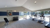 Located at 9015 10 Mile Road in Knoxville, Tennessee, this single-floor fitness center is designed to provide a bright, welcoming environment for exercise and wellness. The space is defined by its soaring vaulted ceilings, which lend an open, airy feel throughout. Large floor-to-ceiling windows and glass doors line one wall, flooding the interior with natural light and offering views of the surrounding outdoor grounds, creating an energizing atmosphere for workouts. The flooring features neutral-toned carpet tiles that add comfort underfoot while maintaining a clean, modern aesthetic. A dedicated free weights area is equipped with a full rack of dumbbells in varying sizes, accompanied by a decorative wall mirror that enhances the sense of space. On the cardio side, the facility includes a treadmill, elliptical trainer, and stationary bike, all positioned to take advantage of the natural light from the expansive windows. The walls are finished in a neutral palette accented by bold navy blue panels, contributing to a focused and motivating workout environment. Recessed lighting supplements the natural illumination for use at any hour. The space is thoughtfully laid out to balance open floor space with functional equipment placement, making it suitable for a variety of fitness routines.