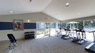 Located at 9015 10 Mile Road in Knoxville, Tennessee, this single-floor fitness center is designed to provide a bright, welcoming environment for exercise and wellness. The space is defined by its soaring vaulted ceilings, which lend an open, airy feel throughout. Large floor-to-ceiling windows and glass doors line one wall, flooding the interior with natural light and offering views of the surrounding outdoor grounds, creating an energizing atmosphere for workouts. The flooring features neutral-toned carpet tiles that add comfort underfoot while maintaining a clean, modern aesthetic. A dedicated free weights area is equipped with a full rack of dumbbells in varying sizes, accompanied by a decorative wall mirror that enhances the sense of space. On the cardio side, the facility includes a treadmill, elliptical trainer, and stationary bike, all positioned to take advantage of the natural light from the expansive windows. The walls are finished in a neutral palette accented by bold navy blue panels, contributing to a focused and motivating workout environment. Recessed lighting supplements the natural illumination for use at any hour. The space is thoughtfully laid out to balance open floor space with functional equipment placement, making it suitable for a variety of fitness routines.