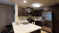 The kitchen is a standout feature of this single-floor space, thoughtfully designed to balance both functionality and aesthetic appeal. Dark espresso-toned cabinetry lines the walls, providing ample storage through upper and lower cabinet configurations. The rich, dark wood tones are complemented by crisp white countertops, creating a striking visual contrast throughout the space.

A generously sized peninsula with a built-in undermount sink serves as the kitchen's centerpiece, offering additional prep space and a natural gathering point. The stainless steel appliance suite includes a French door refrigerator with water and ice dispenser, a gas range with oven, and an over-the-range microwave, all contributing to a cohesive, modern aesthetic.

A textured stone tile backsplash runs beneath the upper cabinets, adding depth and visual interest between the countertops and cabinetry. Recessed and flush-mount ceiling lighting provide ample illumination throughout, keeping the workspace bright and functional. The overall design presents a clean, contemporary kitchen well-suited for both everyday cooking and entertaining.